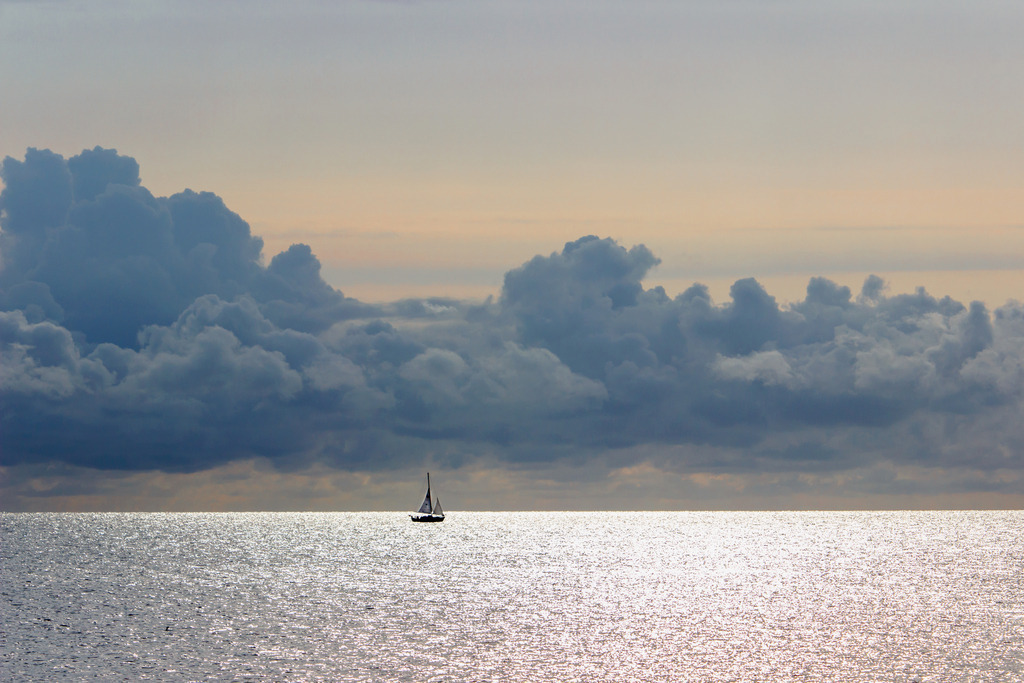 Wandbild: Segelboot im Morgenrot vor malerischen Wolken | Dieses Wandbild im Querformat ein Segelboot in schöner Morgenstimmung vor malerischen Wolken. Der Himmel ist dezent rötlich. Auf dem Meer ergeben sich zahlreiche Lichtreflexionen. Durch die morgendliche Lichtstimmung ergibt sich ein schöner natürlicher Sepiaton. - Realisiert mit Pictrs.com