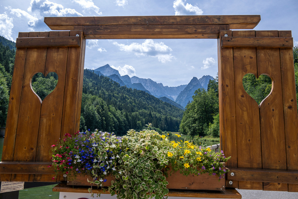 Herzlich willkommen in den Alpen: Blick vom Riessersee auf die Alpspitze | Ein uriger Holzrahmen mit geschnitzten Herzen und bunten, blühenden Sommerblumen setzt die alpine Natur perfekt in Szene. Der Blick vom idyllischen Riessersee gibt eine traumhafte Aussicht auf die dicht bewaldeten Hänge und die imposante Silhouette der Alpspitze frei, die sich stolz gen Himmel streckt. Die Kombination aus traditioneller Handwerkskunst und imposanter Bergwelt vermittelt Heimatgefühl, Gemütlichkeit und die pure Magie eines Sommertages in den Bergen. Ein Motiv, das sofort Herzklopfen und Wanderlust weckt. - Realisiert mit Pictrs.com
