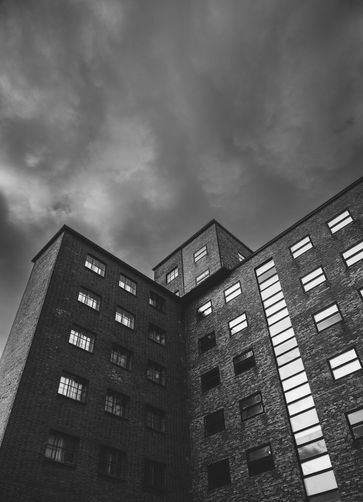 Beneath the Weight | Towering walls of brick and steel rise beneath a sky that feels almost unbearably heavy. The architecture stands firm, yet the atmosphere above suggests tension, pressure, and the quiet force of something far greater.The dark tones and upward perspective create a sense of confinement, while the windows remain silent — holding stories that are never fully revealed. This image captures the fragile balance between permanence and pressure, where the city feels both powerful and vulnerable at once. - Realisiert mit Pictrs.com