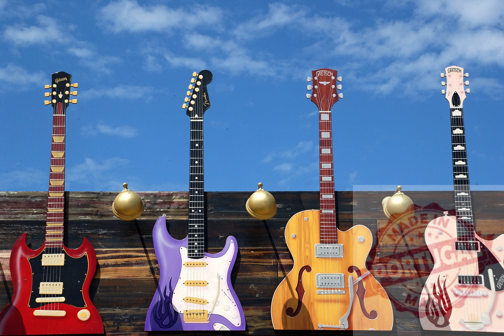Four colorful electric guitars hanging on a wooden wall under a blue sky | Four colorful electric guitars are hanging on a wooden wall under a blue sky, creating a vibrant and musical scene - Realized with Pictrs.com