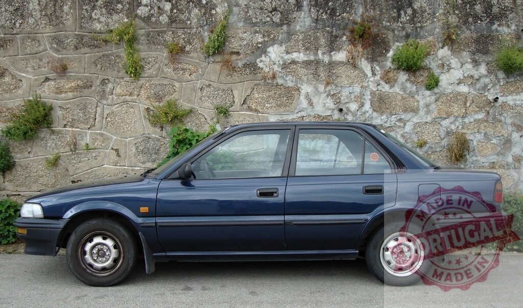 Dark blue Toyota Corolla E90 parked near stone wall with growing plants | Side view of a dark blue Toyota Corolla E90 sedan parked on asphalt road near an old stone wall with some growing plants - Realisiert mit Pictrs.com