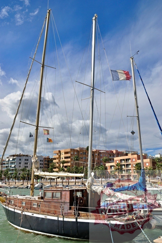 Sailboat moored in harbor with city and national flags | Sailboat moored in marina with building architecture and flags under a bright sky - Realized with Pictrs.com