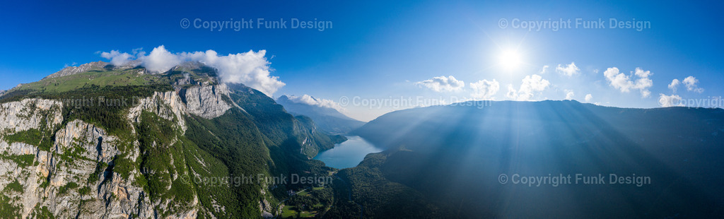 Sonnenstrahlen über den Bergen – Trentino, Italien | Ein weites Drohnenpanorama zeigt eine mächtige Felswand und dahinter ein Tal mit See, während die Sonne rechts im Bild starke Lichtstrahlen über die Berge wirft. Die Szene wirkt episch, frei und klar und eignet sich perfekt als großes Panorama-Wandbild.