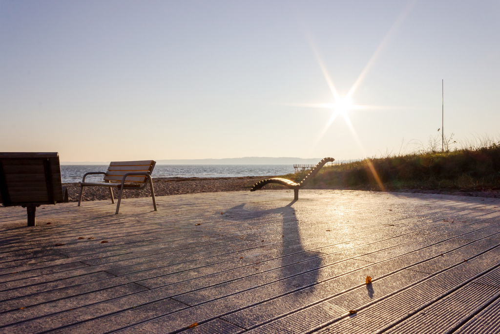 Wandbild: Morgenstimmung am Strand in Eckernförde | Dieses Wandbild im Querformat zeigt die morgendliche Sonne am Strand in Eckernförde. Die Sonnenstrahlen sind sehr gut zu erkennen. Im Vordergrund sind Bänke zu sehen.  - Realisiert mit Pictrs.com