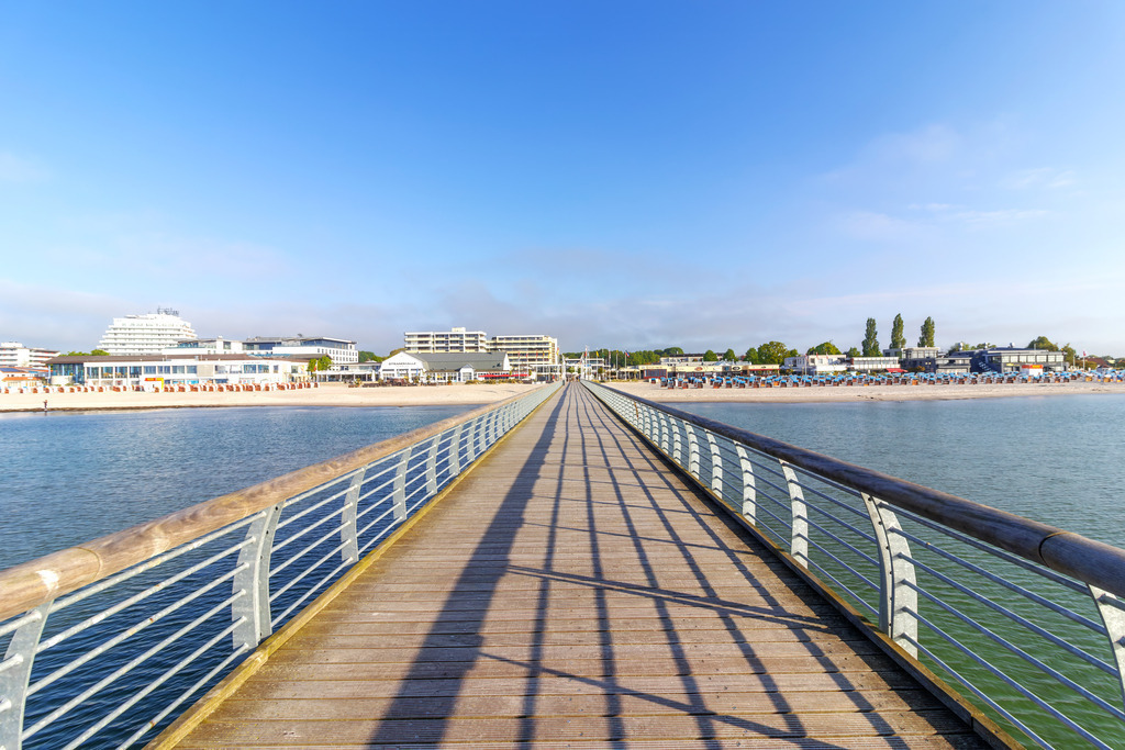 XXL Wandbild Grömitz – Blick von der Seebrücke auf den Sandstrand | Dieses maritime XXL Wandbild zeigt den geradlinigen Blick entlang der Seebrücke von Grömitz – eine Perspektive, die Ruhe und Weite vermittelt. Die Holzplanken der Brücke führen den Blick direkt auf den Strand, wo bunte Strandkörbe aufgereiht stehen und die Küstenkulisse prägen. Rechts und links wird die Brücke von Metallgeländern eingefasst, die die klare Linienführung unterstreichen. Im Hintergrund erheben sich moderne Gebäude, darunter Hotels und Ferienanlagen, eingebettet in grüne Baumgruppen. Der Himmel ist leicht bewölkt und verleiht dem Motiv eine freundliche, offene Stimmung. Dieses Bild bringt die entspannte Eleganz eines Ostseetages direkt an die Wand und eignet sich ideal für maritime Wohnkonzepte.  - Realisiert mit Pictrs.com
