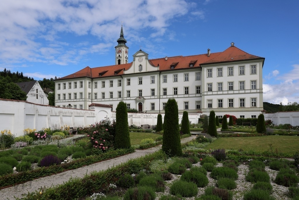 Prälatengarten am Kloster Schäftlarn | Das Foto zeigt den gepflegten Prälatengarten des Kloster Schäftlarn. Die Anlage ist durch klare Formen, symmetrische Wege und sorgfältig angelegte Beete geprägt. Üppige Blumenrabatten, niedrige Hecken und kunstvoll geschnittene Sträucher verleihen dem Garten eine elegante, barock inspirierte Ausstrahlung.Zwischen den Beeten führen gerade Wege durch die Anlage und lenken den Blick auf die harmonische Gesamtgestaltung. Im Hintergrund erhebt sich die historische Klosteranlage mit ihrer beeindruckenden Architektur, die den Garten als würdigen Rahmen ergänzt. Das frische Grün der Pflanzen bildet einen reizvollen Kontrast zu den hellen Fassaden und den warmen Farbtönen der Gebäude.Das Sonnenlicht hebt die feinen Strukturen der Bepflanzung hervor und lässt die Farben der Blüten lebendig erscheinen. Die Atmosphäre des Fotos wirkt ruhig, geordnet und besinnlich. Der Prälatengarten vermittelt die Verbindung von klösterlicher Tradition, gärtnerischer Kunst und der Schönheit der Natur und lädt zum Verweilen und Genießen ein. - Realisiert mit Pictrs.com