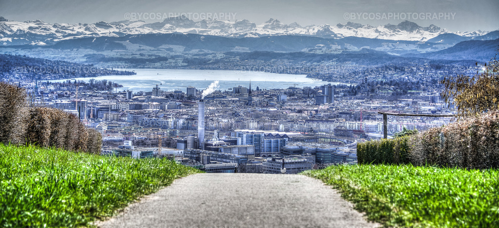 2 | Weitläufiger Panoramablick über die Stadt Zürich zum Zürichsee und zu den schneebedeckten Schweizer Alpen. Der Weg im Vordergrund führt den Blick durch die grüne Landschaft bis zur Stadt, zum See und zum eindrucksvollen Bergpanorama. Ideal als Wandbild, hochwertiger Fotoprint oder Fotoprodukt. - Realisiert mit Pictrs.com