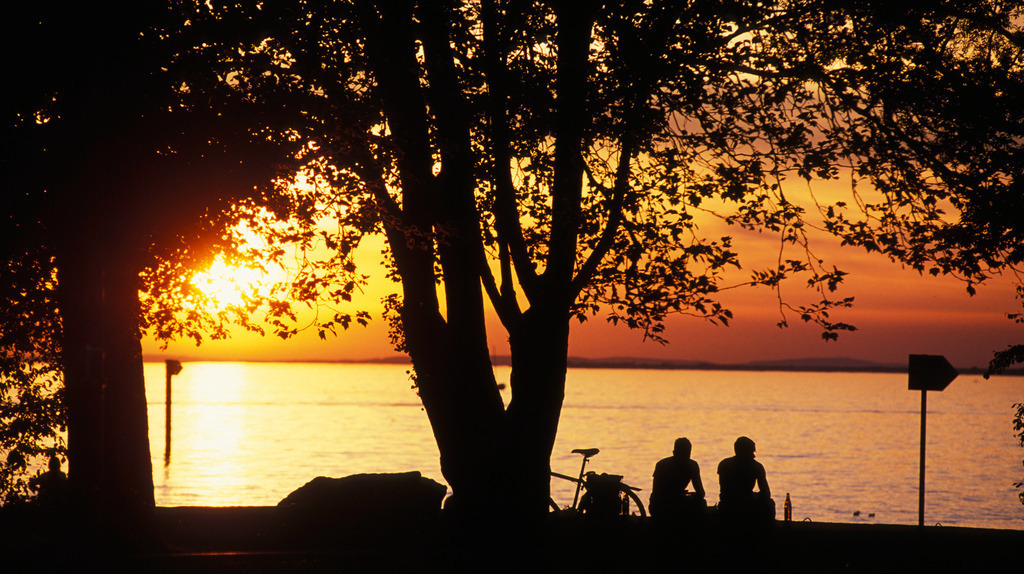 Freunde am See | Bregenz, Austria: Zwei Freunde mit Fahrrad sitzen am Ufer des Bodensees im Abendrot. - Realisiert mit Pictrs.com