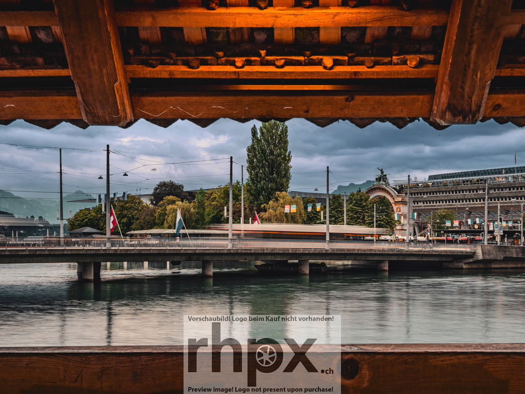 Bahnhofblick durch die Kapellbrücke | Rahmen. Zeit. Dynamik.Das historische Gebälk der Kapellbrücke bildet einen natürlichen Rahmen für das pulsierende Leben rund um den Luzerner Bahnhof. Die Langzeitbelichtung lässt den vorbeiziehenden Verkehr auf der Seebrücke zu weichen Spuren verschmelzen und fängt die städtische Energie in einem ruhigen Moment ein.Wähle unter "Produktauswahl" dein Wunschformat: Vom klassischen Wandbild über Puzzle &amp; Tassen bis zum digitalen Download (z.B. als Handy-Hintergrund). - Realisiert mit Pictrs.com
