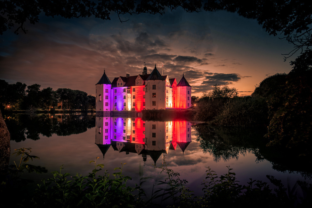 Schloss Glücksburg | Das Bild zeigt das majestätische Schloss in Glücksburg, welches Nachts mit den Farben von Schleswig-Holstein angestrahlt wird. Die warmen Farben der Beleuchtung lassen das Schloss in einem atemberaubenden Licht erstrahlen und betonen seine prächtigen Türme und Fassaden.
Die sanfte Beleuchtung reflektiert sich im Schlossteich und schafft eine zauberhafte Atmosphäre, die den Betrachter in eine Welt voller Magie und Schönheit entführt. Die ruhige Wasseroberfläche lässt das Schloss wie in einem Spiegelbild erscheinen und erzeugt eine fast surreal wirkende Stimmung. Die Szenerie lädt zum Träumen und Verweilen ein. Es erinnert an die Geschichte und Bedeutung des Schlosses in Glücksburg als bedeutendes Kultur- und Geschichtsdenkmal der Region Schleswig-Holstein.