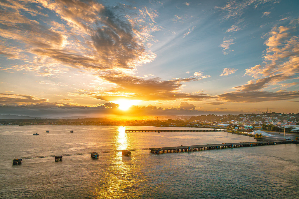 Stimmungsvoller Sonnenuntergang über die Küstenstadt Puerto Limón in Costa Rica | Das ausgewählte Bild zeigt einen stimmungsvollen Sonnenuntergang über der Küstenstadt Puerto Limón in Costa Rica.Blick auf die Bucht und die Hafenanlagen bzw. Piers von Puerto Limón an der Karibikküste Costa Ricas.Die Szenerie fängt die goldene Stunde ein, bei der die tiefstehende Sonne das Wolkenband in leuchtenden Orange- und Gelbtönen erstrahlen lässt und ausgeprägte Lichtstrahlen (Krepuscularstrahlen) durch die Wolkendecke brechen. Im Vordergrund erstreckt sich eine lange Steganlage über das ruhige Meer - Realisiert mit Pictrs.com