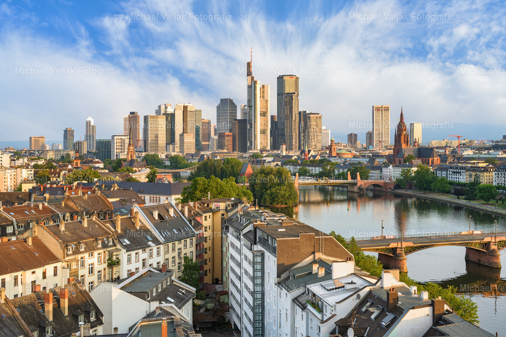 Frankfurt in der Morgensonne | Der Blick über die Dächer Frankfurts zeigt eine Stadt in den ersten Momenten des Tages. Kurz nach Sonnenaufgang liegt ein warmes Licht über den Gebäuden, das die Fassaden sanft hervorhebt. Die Skyline mit dem Bankenviertel hebt sich klar am Horizont ab – modern, strukturiert und ruhig.Die Sonnenstrahlen spiegeln sich in den Glasflächen der Hochhäuser, während die Stadt langsam erwacht. Es ist ein alltäglicher Moment, aber mit eigener Atmosphäre: still, konzentriert und weit entfernt vom Trubel des Tages.Ein Bild, das den Kontrast zwischen Wohnvierteln und Wirtschaftszentrum aufzeigt – unaufgeregt und präzise. - Realisiert mit Pictrs.com