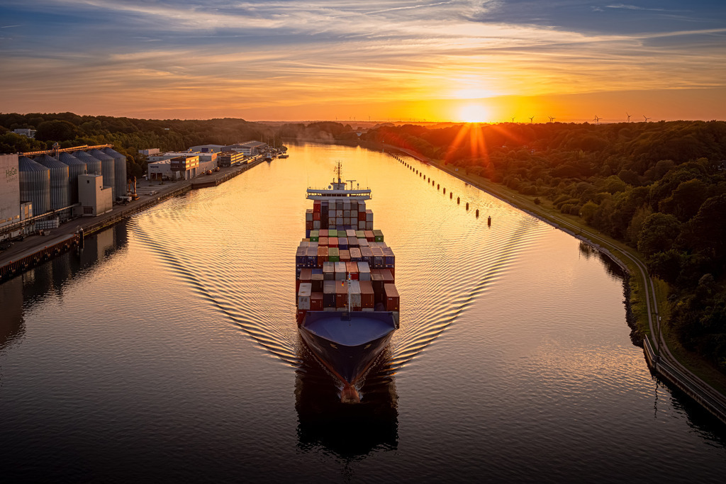 Sonnenuntergang am Nord Ostsee Kanal | Auf dem Foto, das von der Holtenauer Hochbrücke aus aufgenommen wurde, erstreckt sich der weitläufige Nord-Ostsee-Kanal vor einem atemberaubenden Sonnenuntergangshimmel. Die Szene strahlt eine friedliche Gelassenheit aus, während die Sonne in warmen, orange-roten Farbtönen hinter dem Horizont versinkt. Ein beeindruckendes Containerschiff dominiert die Bildmitte.
Die gesamte Szenerie strahlt eine ruhige, fast magische Atmosphäre aus, die die Schönheit der Natur und die menschliche Ingenieurskunst in harmonischem Einklang zeigt. Dieses Foto fängt den Zauber des Nord-Ostsee-Kanals bei Sonnenuntergang ein.
