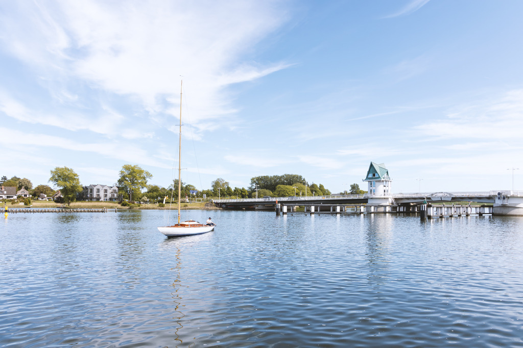 Wandbild: Helles Hafenbild aus Kappeln – Segler vor der Schleibrücke | Dieses Wandbild zeigt eine ruhige Hafenszene in Kappeln, eingefangen in einem klaren, hellen Licht, das die weite Wasserfläche und die freundliche Atmosphäre der Schlei betont. Ein Segler liegt still im Hafen, während die Schleibrücke mit ihrem markanten Turm dezent im Hintergrund erscheint und dem Motiv eine norddeutsche Note verleiht. Die umliegenden Gebäude und Bäume fügen sich harmonisch in die Szenerie ein und unterstreichen die maritime Gelassenheit dieses Moments. Ein Premium‑Wandbild, das mit Helligkeit, Weite und norddeutschem Flair moderne Wohnräume stilvoll ergänzt.