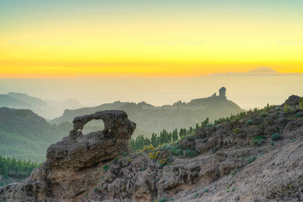 Blick vom Pico de las Nieves auf Gran Canaria | Nach Sonnenuntergang liegt eine besondere Stille über der Bergwelt Gran Canarias. Im Vordergrund steht ein natürlicher Steinbogen aus dunklem Vulkangestein, rau und bizarr geformt durch Wind und Zeit. Dahinter breitet sich die silhouettenhafte Landschaft aus – mit dem markanten Roque Nublo als vertrautem Orientierungspunkt. In der Ferne erhebt sich der Teide auf Teneriffa aus einem ruhigen Wolkenmeer. - Realisiert mit Pictrs.com
