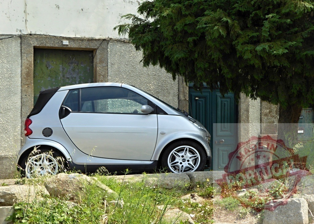 Smart car parked on rough urban terrain | Silver Smart ForTwo compact car parked on rocky ground next to an old building and tree - Realized with Pictrs.com