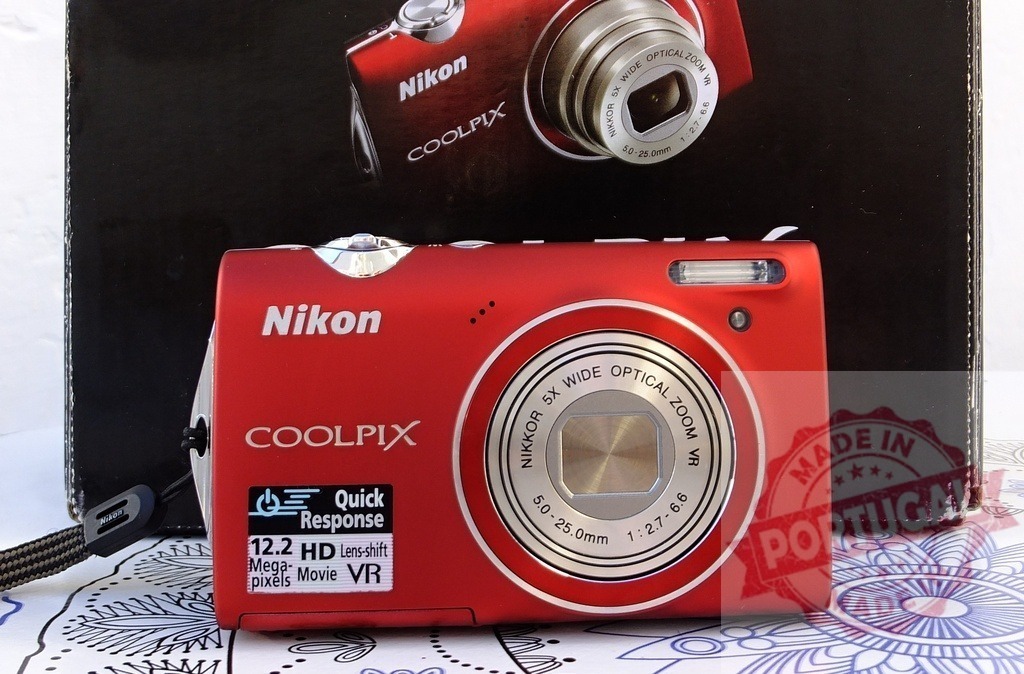 Red Nikon Coolpix digital camera on white background | Red Nikon Coolpix point and shoot camera sitting on a patterned surface with its product box behind - Realisiert mit Pictrs.com