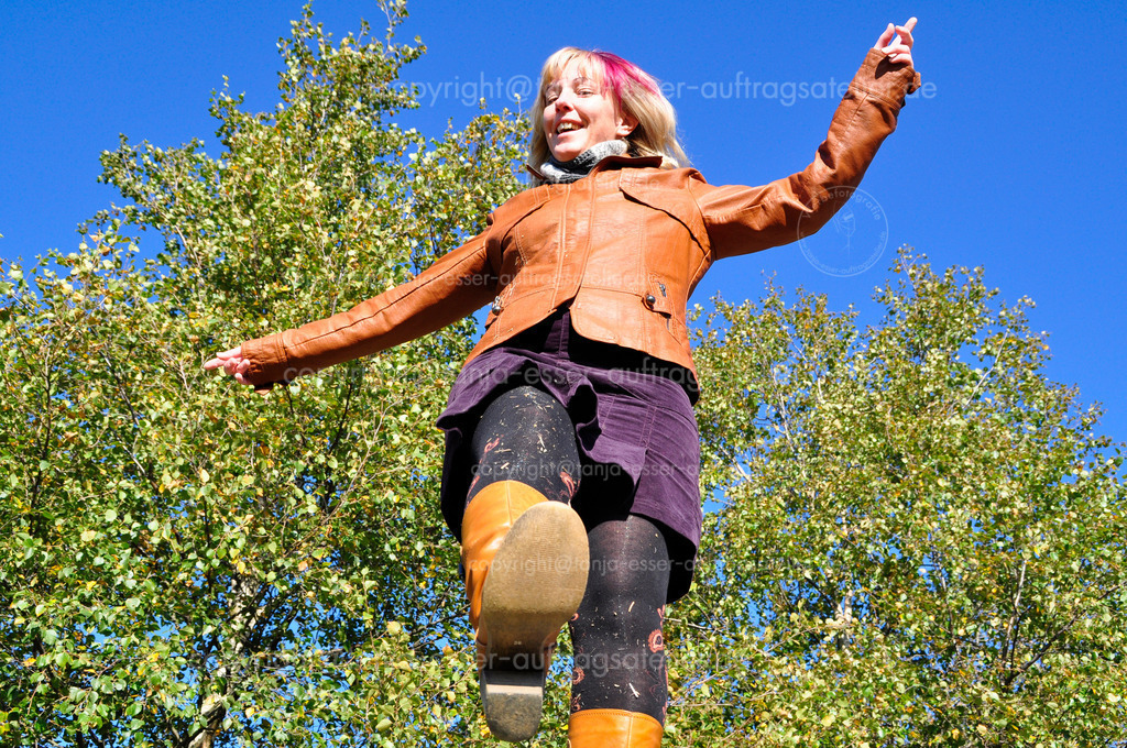 Autumn fun up view | Herbstspaß. Eine blonde Frau Ende 30 klettert im schicken Outfit über Siloballen. Lederjacke und Lederstiefel. Sie hat blonde Haare und eine pinke Haarsträhne. Sie lacht. Froschperspektive.