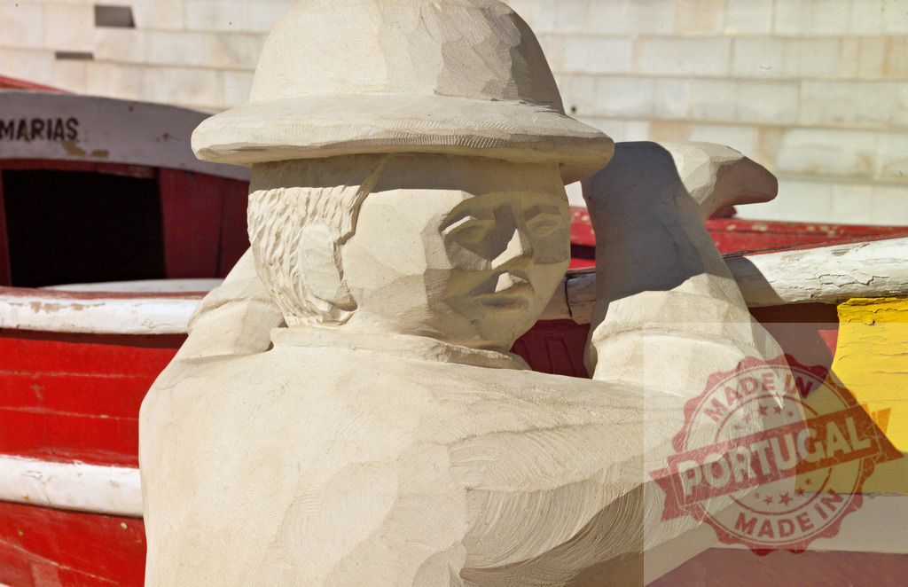 Stone fisherman sculpture with boats in background | Stone sculpture of a fisherman with a hat, standing near red and yellow boats - Realisiert mit Pictrs.com