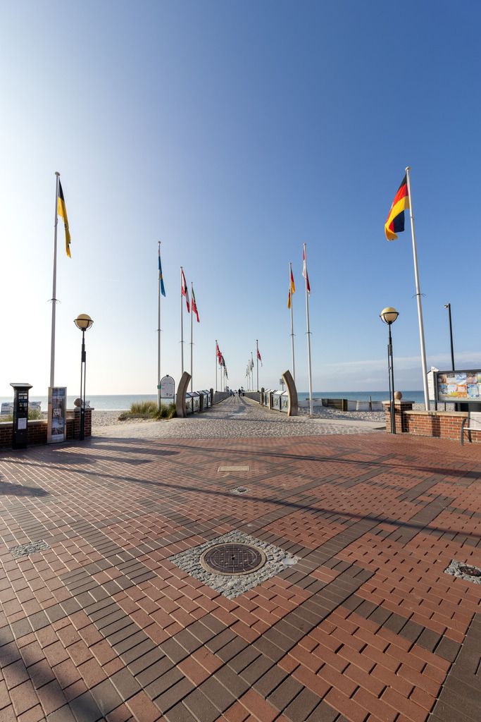 Wandbild: Küstenflair – Grömitzer Seebrücke mit Flaggen | Dieses maritime Wandbild zeigt die gepflegte Promenade von Grömitz, die direkt zur Seebrücke führt. Der Weg ist mit roten Pflastersteinen ausgelegt und wird von modernen Straßenlaternen und Infotafeln gesäumt. Zahlreiche Fahnenmasten mit internationalen Flaggen – darunter auch die deutsche – verleihen dem Ort ein weltoffenes, einladendes Flair. Die Seebrücke erstreckt sich geradewegs ins ruhige Meer, das unter dem klaren blauen Himmel glitzert. Die Komposition wirkt aufgeräumt und symmetrisch, ideal für Liebhaber maritimer Architektur und Ostseeatmosphäre. Dieses Motiv eignet sich hervorragend als Wandbild für maritime Wohnkonzepte – ob als Leinwandbild, Acrylglasbild, Alu-Dibond FineArt Print oder als Akustikbild. Ein stilvoller Blickfang für Zuhause, Büro oder Ferienwohnung. - Realisiert mit Pictrs.com