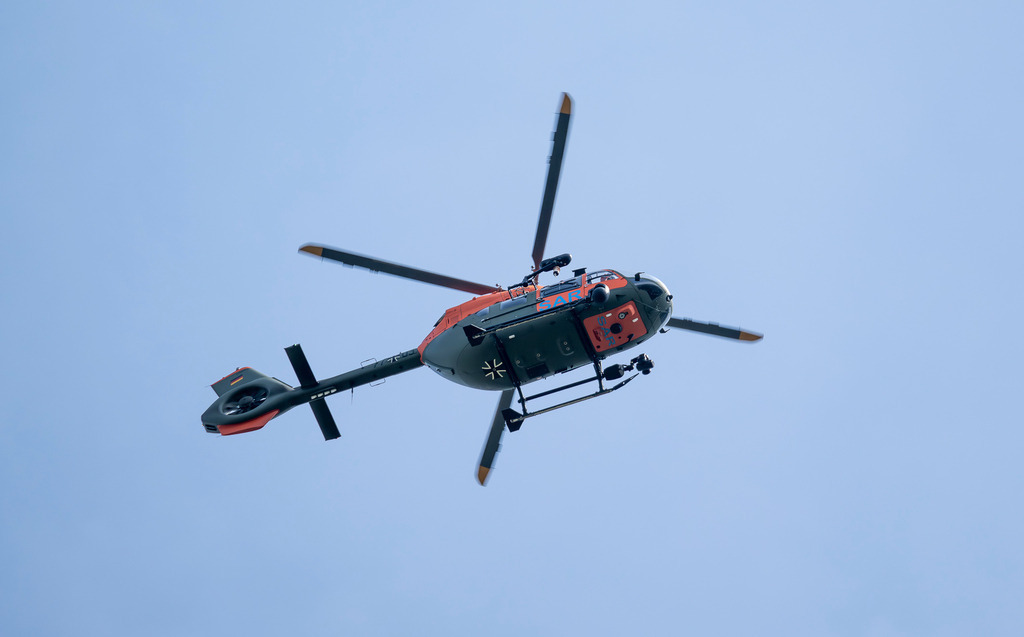 Airbus H145 LUH SAR Hubschrauber | Die Aufnahme zeigt einen Airbus H145 LUH SAR Hubschrauber der Bundeswehr in der Untersicht gegen einen klaren, blauen Himmel. Der Helikopter ist in olivgrün und leuchtendem Orange (Dayglow) lackiert, trägt die Aufschrift „SAR“ (Search and Rescue) sowie das Eiserne Kreuz und verfügt über einen markanten Fenestron-Heckrotor. - Realisiert mit Pictrs.com