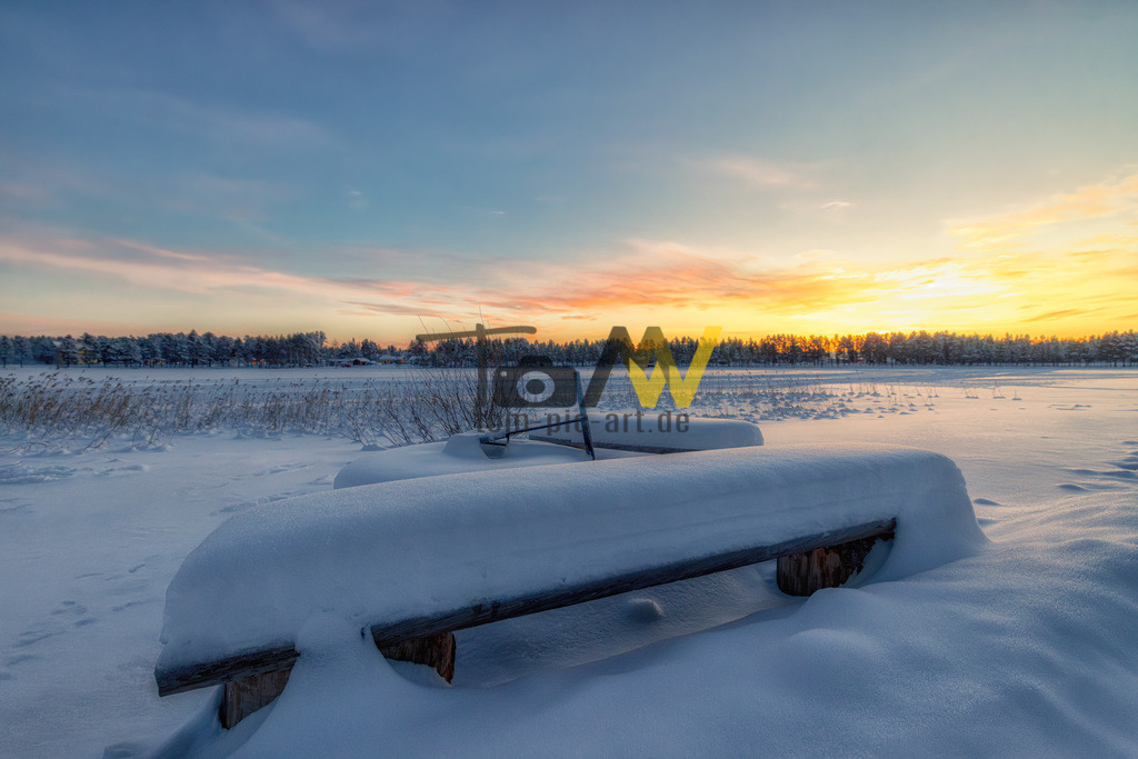 Eine Sitzbank in Schweden mit viel Schnee bei Sonnenaufgang | Eine verschneite Winterlandschaft bei Sonnenaufgang.Im Vordergrund ist eine Holzbank zu sehen, die vollständig mit Schnee bedeckt ist.Im Hintergrund erstreckt sich eine weite, schneebedeckte Ebene, die von einem Waldsaum begrenzt wird.Der Himmel zeigt einen farbenfrohen Sonnenuntergang mit orangefarbenen, gelben und blauen Tönen. - Realisiert mit Pictrs.com