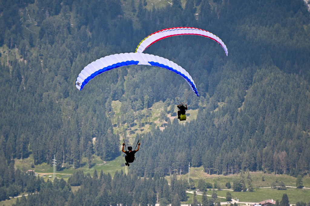 Freiheit im Doppelpack: Gleitschirmduo über den Alpen | Zwei Gleitschirme, einer mit leuchtend blauen und der andere mit rot-weißen Akzenten, schweben majestätisch über einem dichten, grünen Bergwald. Die Aufnahme fängt einen Moment purer Schwerelosigkeit und Freiheit ein. Die Silhouetten der Piloten sind deutlich zu erkennen, wie sie die Lüfte bezwingen und die atemberaubende Alpenkulisse aus der Vogelperspektive genießen. Dieses Bild weckt Sehnsucht nach Abenteuer, Frieden und der grenzenlosen Weite der Natur. Es symbolisiert den menschlichen Traum vom Fliegen und die Harmonie zwischen Mensch und Natur. - Realisiert mit Pictrs.com