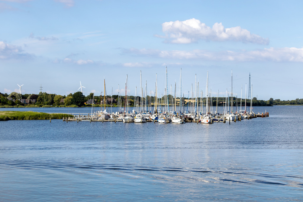 Wandbild: Yachthafen in Lindaunis | Dieses Wandbild im Querformat zeigt den Yachthafen in Lindaunis. Die Segelboote im Hafen spiegeln sich leicht auf der Schlei. Im Vordergrund sind einige Wellen zu sehen. Am blauen Himmel befinden sich einige sommerliche Wolken. - Realisiert mit Pictrs.com
