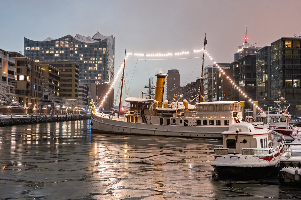 Wintermärchen im Traditionshafen | Der historische Staatsdampfer Schaarhörn an seinem Liegeplatz im Hamburger Sandtorhafen. Die Aufnahme zeigt das beleuchtete Museumsschiff bei Eisgang im Hafenbecken.Im Hintergrund bildet die Glasfassade der Elbphilharmonie den Kontrast zur klassischen Linienführung des Dampfers aus dem Jahr 1908. - Realisiert mit Pictrs.com