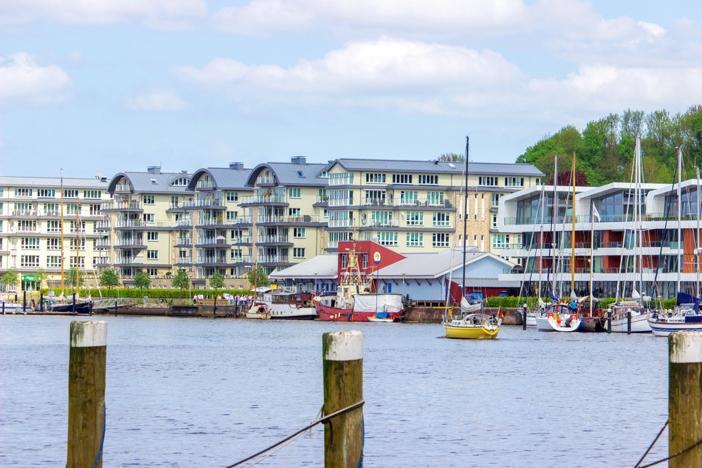 Wandbild: Flensburger Hafen | Dieses Wandbild im Querformat zeigt den Hafen in Flensburg. Auf der anderen Uferseite sind Gebäude zu sehen. Im Vordergrund befinden sich Pfähle. Am blauen Himmel befinden sich einige Wolken.  - Realisiert mit Pictrs.com