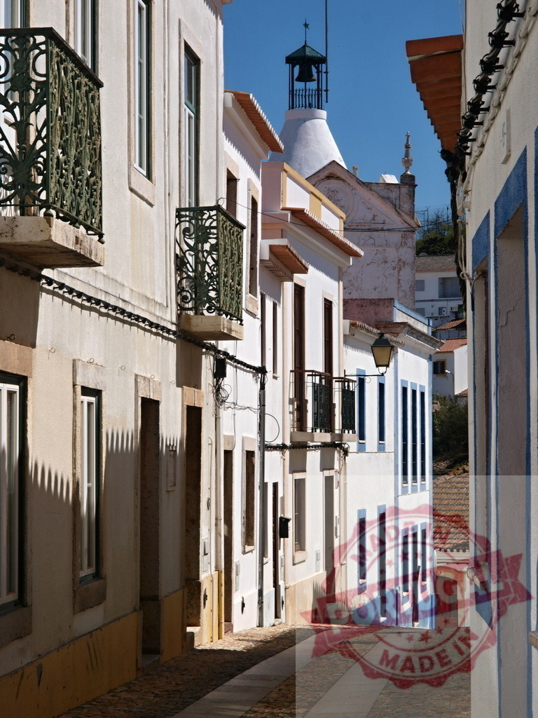 Narrow alley with whitewashed houses and bell tower in Portugal | Charming Portuguese alley featuring traditional whitewashed buildings with balconies and a bell tower under a clear sky - Realisiert mit Pictrs.com