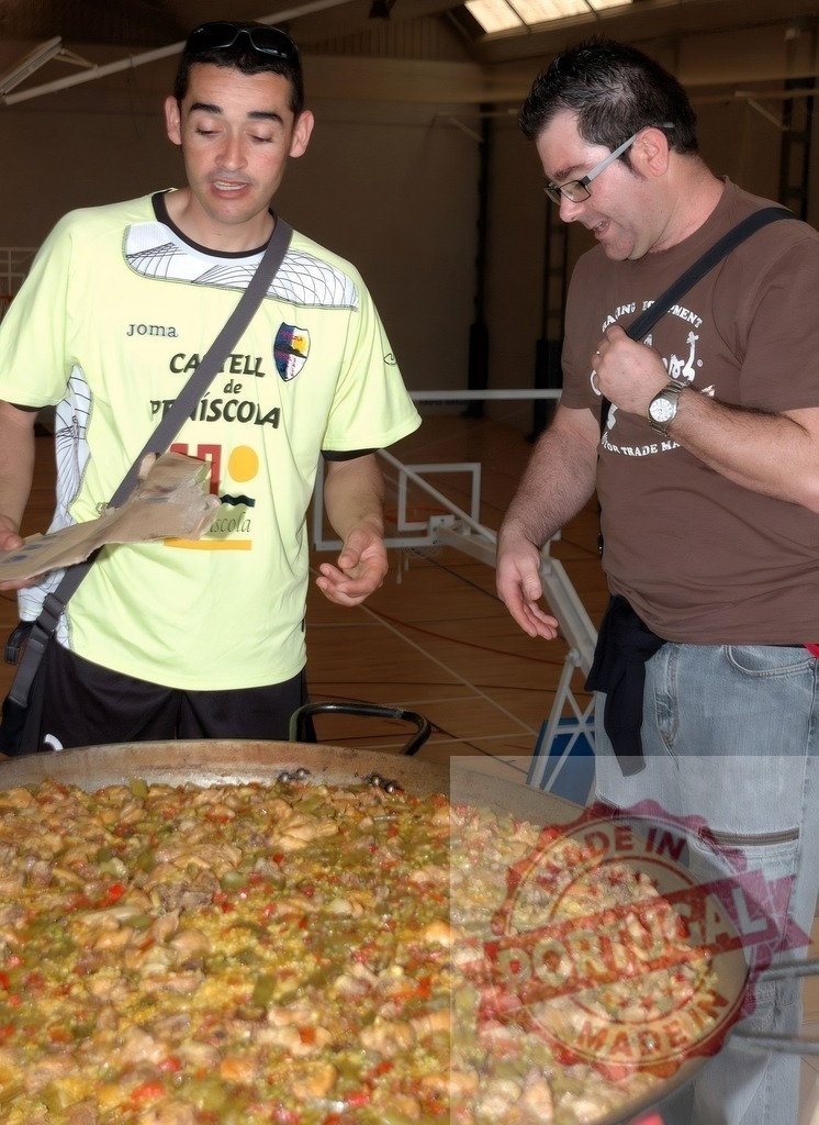 Men enjoying paella at community sports event | Two men are admiring a large paella at a community gathering in a sports hall - Realized with Pictrs.com
