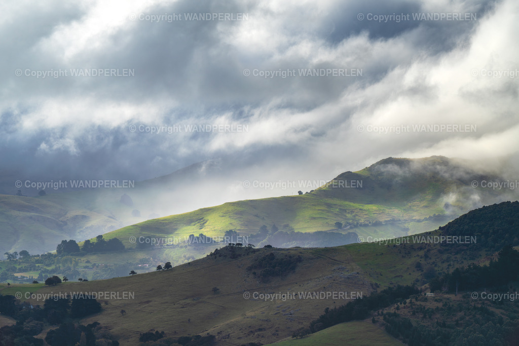Banks Peninsula bei stürmischem Wetter | Eine weite Ansicht welliger, grüner Hügel wird teilweise von einer dichten Schicht niedriger Wolken, was eine stimmungsvolle, atmosphärische Szene erzeugt. Sonnenlicht dringt an einzelnen Stellen durch, beleuchtet Teile der Landschaft und wirft tiefe Schatten in Täler und Hänge. Das Bild zeigt einen Moment dynamischen Wetters, die Wolken ziehen schnell über die Landschaft. - Realisiert mit Pictrs.com