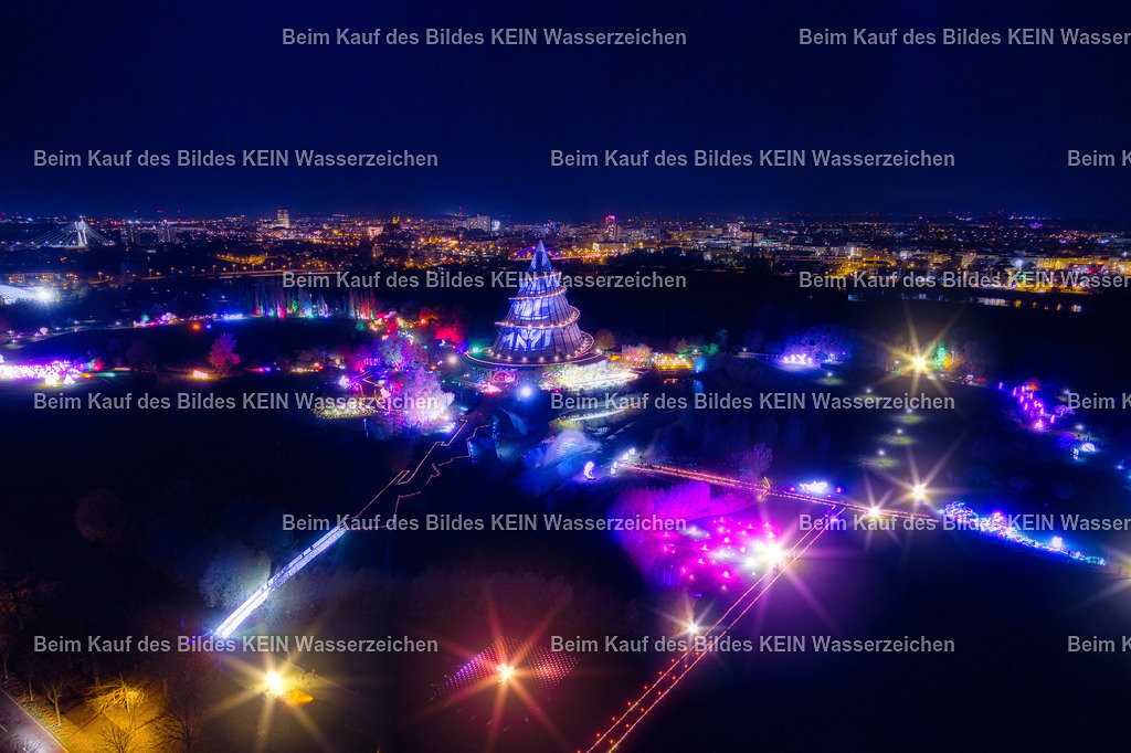 lumagica_Magdeburg_Jahrtausendturm_Nachts_0012 | Das Luftbild zeigt den Jahrtausendturm im Elbauenpark Magdeburg während der Lichtershow Lumagica aus einer Drohnenperspektive. Die farbenfrohen Lichtinstallationen setzen die markante Holzkonstruktion eindrucksvoll in Szene und lassen die außergewöhnliche Architektur des Bauwerks aus der Vogelperspektive besonders gut erkennen. Die nächtliche Beleuchtung hebt die geometrische Form des Turms hervor und macht ihn zum zentralen Blickfang der Aufnahme.Der Jahrtausendturm wurde zur Bundesgartenschau 1999 errichtet und ist mit einer Höhe von 60 Metern der höchste hölzerne Aussichtsturm seiner Art weltweit. Die freitragende Konstruktion aus Brettschichtholz gilt als Meisterleistung des modernen Holzingenieurbaus. Im Inneren befinden sich auf fünf Ebenen Ausstellungen zur Entwicklung von Technik und Wissenschaft über einen Zeitraum von rund 6.000 Jahren.Die Drohnenaufnahme vermittelt einen außergewöhnlichen Blick auf die Kombination aus moderner Architektur und aufwendiger Lichtinszenierung. Während der Lumagica-Lichtershow verwandeln tausende Lichtquellen den Elbauenpark in ein farbenfrohes Gesamtkunstwerk und setzen den Jahrtausendturm als eines der markantesten Bauwerke Magdeburgs eindrucksvoll in Szene. - Realisiert mit Pictrs.com