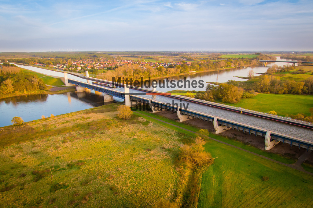 Trogbrücke-5843 | Das Luftbild zeigt die Trogbrücke des Wasserstraßenkreuzes Magdeburg aus einer Drohnenperspektive. Das Bauwerk wird vom warmen orangefarbenen Licht geprägt und hebt sich eindrucksvoll von der umgebenden Landschaft ab. Aus der Vogelperspektive werden die klaren Linien der Brückenkonstruktion und die außergewöhnlichen Dimensionen der Wasserstraße besonders deutlich sichtbar.Die Trogbrücke wurde 2003 eröffnet und ist mit einer Gesamtlänge von 918 Metern die längste Kanalbrücke Europas. Der wassergefüllte Brückentrog führt den Mittellandkanal über die Elbe und ermöglicht der Binnenschifffahrt eine durchgehende Verbindung zwischen den west- und ostdeutschen Wasserstraßen. Der Trog ist 34 Meter breit und rund 4,25 Meter tief, sodass auch große Gütermotorschiffe und Schubverbände die Brücke sicher passieren können. Für den Bau wurden rund 24.000 Tonnen Stahl und etwa 68.000 Kubikmeter Beton verwendet.Die Drohnenaufnahme verdeutlicht eindrucksvoll die Dimensionen und die technische Leistungsfähigkeit dieses außergewöhnlichen Ingenieurbauwerks. Die Kombination aus moderner Stahlbaukunst, innovativer Wasserbautechnik und dem warmen Licht macht die Trogbrücke zu einem der bedeutendsten technischen Wahrzeichen des Wasserstraßenkreuzes Magdeburg. - Realisiert mit Pictrs.com