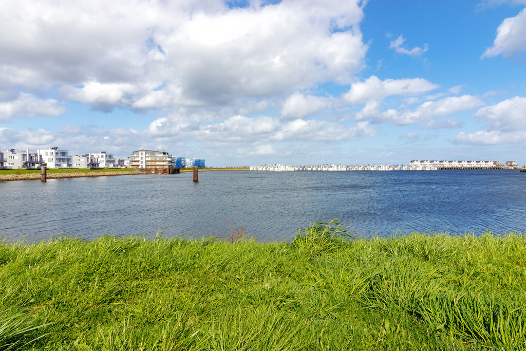 Wandbild: Frühling in Olpenitz | Dieses Wandbild im Querformat zeigt den Blick von der Promenade auf das Feriengebiet in Olpenitz. Im Vordergrund ist der mit Gräsern bewachsene Uferbereich zu sehen. Am blauen Himmel sind schöne frühlingshafte Wolken zu sehen. - Realisiert mit Pictrs.com