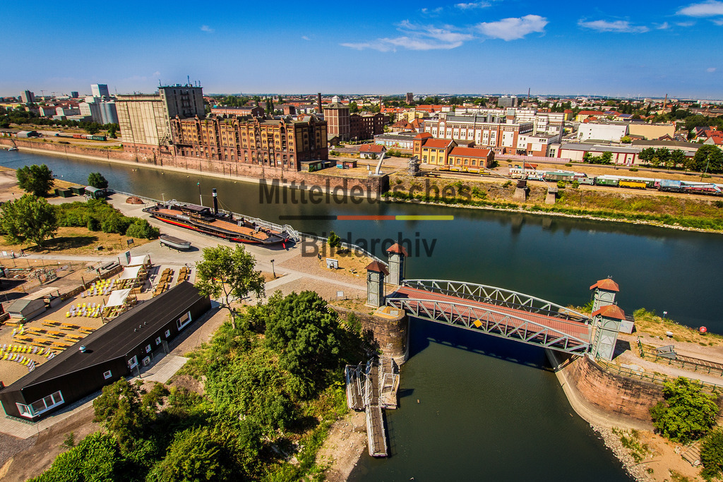 Wissenschaftshafen_Hubbrücke_Cafe-Treibgut_Röstfein-3248 | Das Foto zeigt die kleine Hubbrücke im Wissenschaftshafen Magdeburg aus einer bodennahen Perspektive. Die kompakte Stahlfachwerkkonstruktion mit ihrem markanten Hubportal vermittelt eindrucksvoll die Ingenieurtechnik historischer Hafenanlagen. Durch die niedrige Kameraposition treten die genieteten Stahlträger, der bewegliche Brückenüberbau und die technischen Details besonders deutlich hervor.Die Hubbrücke wurde Anfang des 20. Jahrhunderts als Teil der Hafeninfrastruktur errichtet und diente dem Anschluss der Gleisanlagen im Handelshafen. Über den elektrischen Hubmechanismus konnte der Brückenüberbau angehoben werden, sodass Schiffe den Hafenarm ungehindert passieren konnten. Hubbrücken dieser Bauart galten damals als besonders platzsparende Lösung, da sie im Gegensatz zu Dreh- oder Klappbrücken nur wenig Raum für ihre Bewegung benötigten. Die genietete Stahlkonstruktion ist ein typisches Beispiel des deutschen Stahlbrückenbaus jener Zeit.Die Aufnahme dokumentiert ein selten erhaltenes technisches Denkmal der Magdeburger Hafen- und Eisenbahngeschichte. Die kleine Hubbrücke erinnert an die enge Verbindung von Schifffahrt und Schienenverkehr und steht bis heute als Zeugnis der leistungsfähigen Hafenlogistik, die Magdeburg über viele Jahrzehnte zu einem bedeutenden Binnenhafen machte. - Realisiert mit Pictrs.com