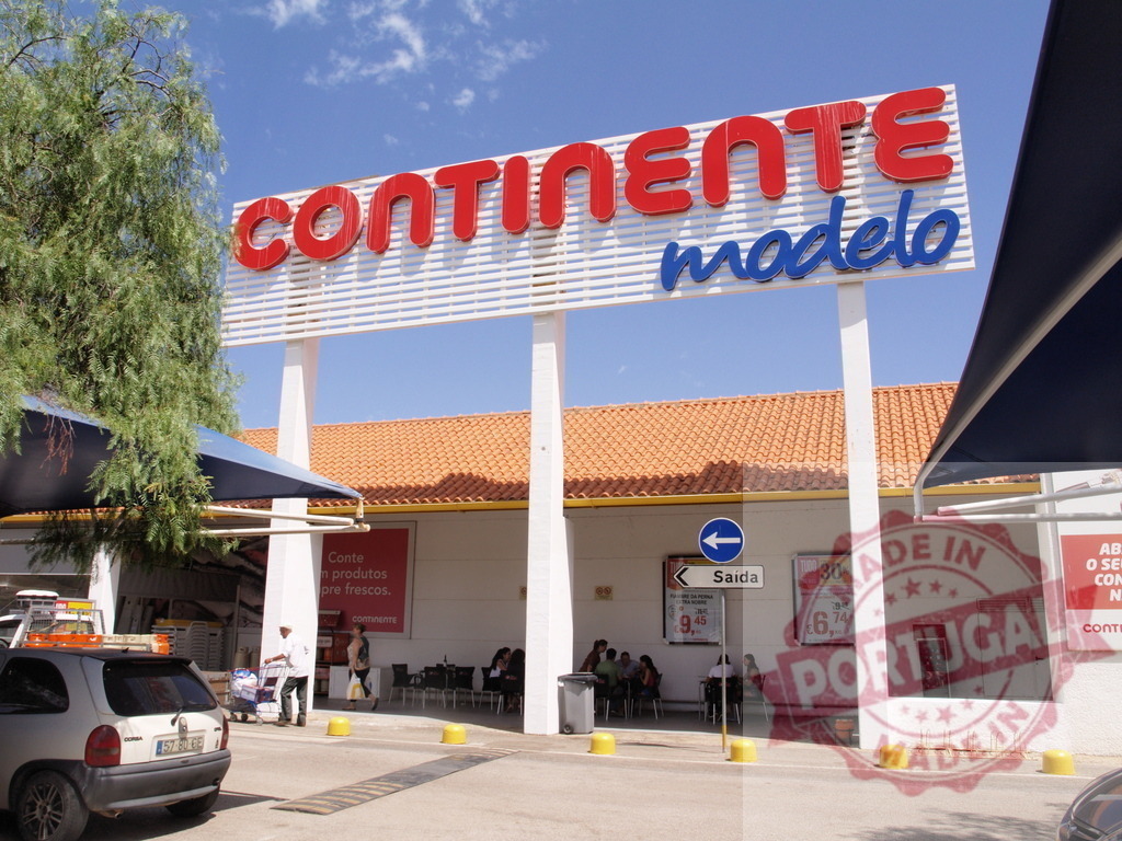 Continente Modelo supermarket exterior entrance on sunny day | Continente Modelo supermarket exterior featuring its prominent sign, people entering and exiting on a sunny day - Realized with Pictrs.com