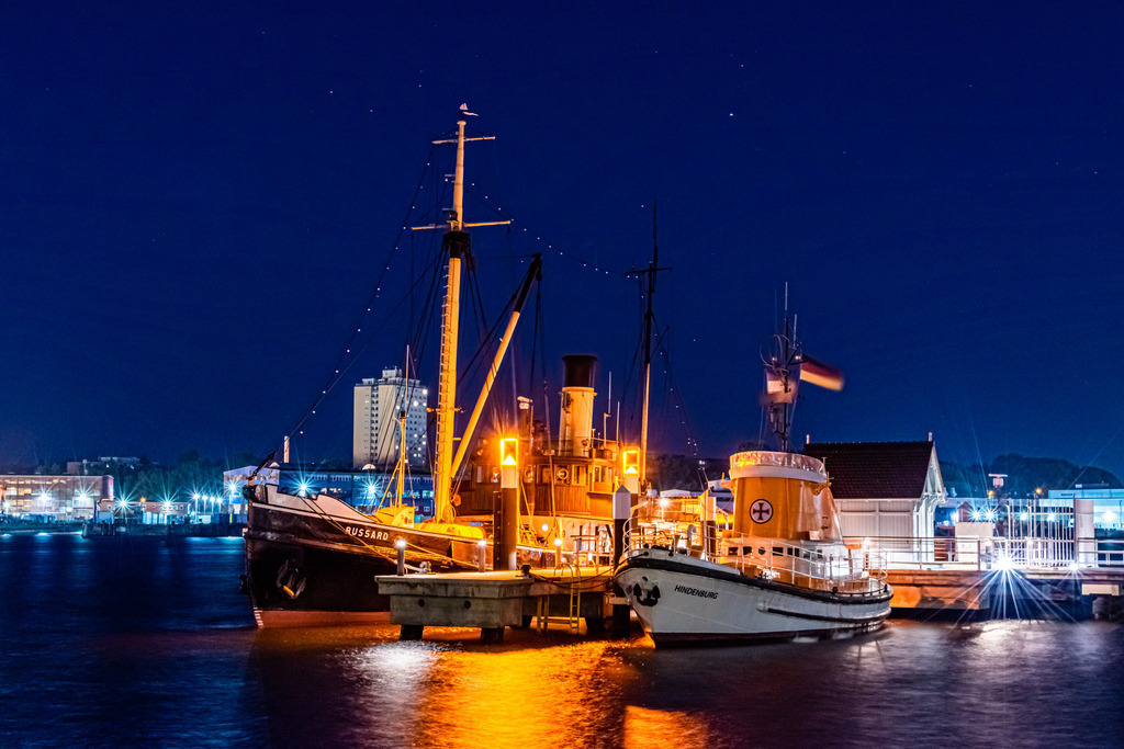 Nachts am Schifffahrtsmuseum - Museumsbrücke | Das Bild zeigt zwei wunderschöne und historische Schiffe, den alten dampfbetriebenen Tonnenleger Bussard und den alten Seenotrettungskreuzer Hindenburg, die an der Kieler Museumsbrücke liegen. Die Schiffe sind ein wahrer Hingucker und erinnern an die maritime Geschichte und Tradition der Stadt Kiel.

Der Tonnenleger Bussard, einst im Dienst der Küstenwache, ist ein beeindruckendes Beispiel für die maritime Technologie des 19. Jahrhunderts und wird durch seine markante Bauweise und seine historischen Details zu einem wahren Erlebnis für jeden Besucher.

Der Seenotrettungskreuzer Hindenburg, der einst im Dienst der Deutschen Gesellschaft zur Rettung Schiffbrüchiger (DGzRS) stand, beeindruckt durch seine Größe und seine kraftvolle Formgebung. Das Schiff erinnert an die unzähligen Rettungsaktionen, die von der DGzRS durchgeführt wurden und werden.

Die Schiffe, die nebeneinander an der Museumsbrücke liegen, bilden eine perfekte Kulisse für das maritime Flair der Stadt Kiel. Eine Szenerie, die den Betrachter auf eine Reise in die Vergangenheit entführt und die Schönheit der maritimen Geschichte und Traditionen der Region zum Ausdruck bringt.