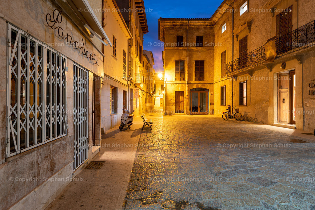 Plaça Vella in Pollença | Blick auf den Plaça Vella (Alter Platz) in Pollença zur blauen Stunde. Links sind die Gitterfenser einer Panaderia (Bäckerei) zu sehen, im Hindergrund steht eine Vespa und ein Fahrrad. - Realisiert mit Pictrs.com