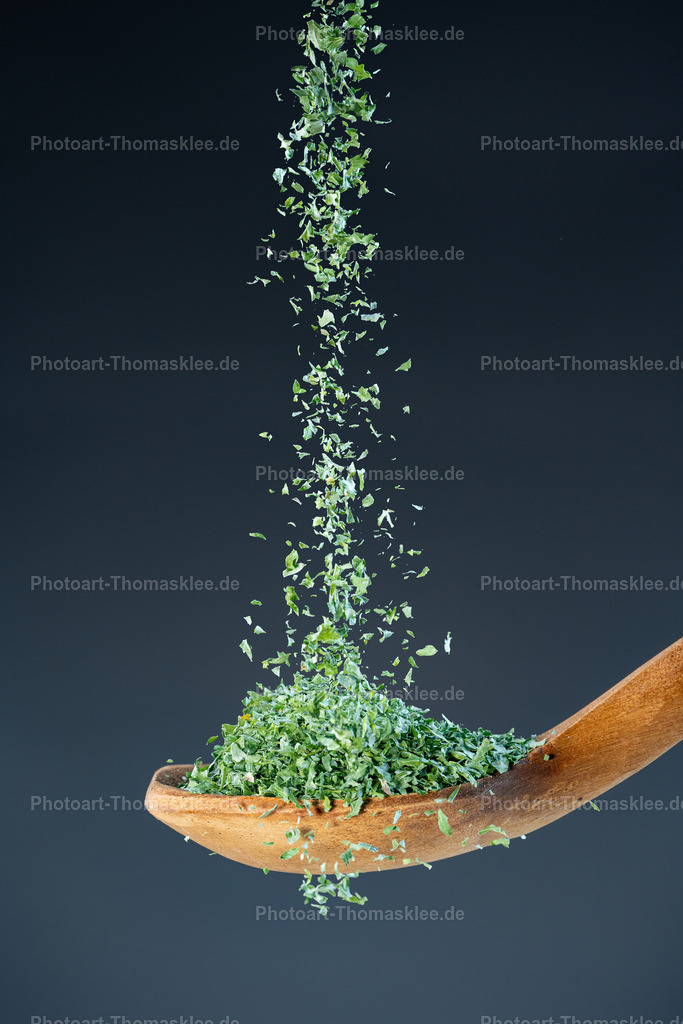Getrocknete Petersilie – Aromatische Kräuter für die feine Küche | Fein zerkrümelte grüne Petersilie fallen in einem lebendigen Strahl aus der Bildmitte herab und sammeln sich in einem naturbelassenen Holzlöffel. Die winzigen Blattstücke wirbeln sanft durcheinander, bevor sie sich zu einem kleinen Hügel in der Löffelmulde formen. Der glatte, matte Grauverlauf im Hintergrund sorgt für eine ruhige, moderne Kulisse und hebt die frischen Grüntöne plastisch hervor.High-Speed-Makrofotografie mit sehr kurzer Verschlusszeit: Jede Kräuterfaser ist gestochen scharf eingefangen, selbst im freien Fall.Geringe Tiefenschärfe: Nur die fallenden Kraut­partikel und der Löffel sind fokussiert, Vorder- und Hintergrund laufen ins sanfte Bokeh über.Diffuses, weiches Licht: Betont die zarte Textur der Kräuter ohne harte Schatten oder grelle Reflexe.Leuchtendes Frühlingsgrün der getrockneten KräuterflockenWarme Honig- bis Nuss­töne im HolzlöffelNeutrales Mittelgrau im Hintergrund für klaren, zeitgemäßen KontrastEmpfohlene Orte zum AufhängenKüche oder Esszimmer – als inspirierender Blickfang, der Frische und Natürlichkeit vermittelt.Feinkostladen oder Kräuterfachgeschäft – um die Vielfalt und Qualität aromatischer Kräuter visuell zu präsentieren. - Realisiert mit Pictrs.com