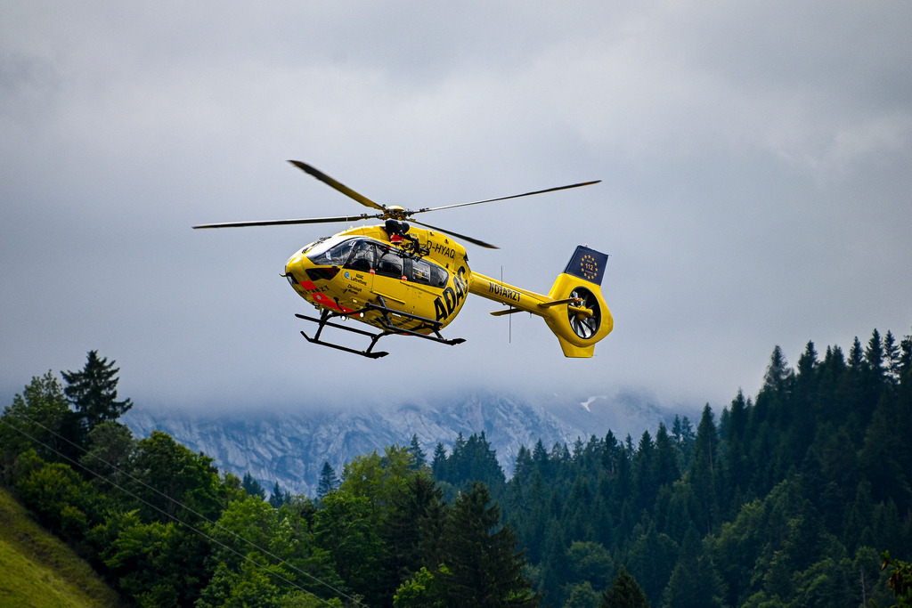 Hoffnung über den Gipfeln | Ein gelber ADAC-Rettungshubschrauber schwebt dynamisch vor einer imposanten Bergkulisse. Während im Hintergrund bewaldete Hänge und wolkenverhangene Felsmassive eine raue alpine Kulisse bilden, sticht die Maschine als leuchtendes Symbol für schnelle Hilfe hervor. Das Bild fängt das perfekte Zusammenspiel aus hochmoderner Technik, menschlichem Einsatz und der Herausforderung der Natur ein. Es strahlt sowohl drängende Dringlichkeit als auch tiefes Vertrauen und Sicherheit aus. - Realisiert mit Pictrs.com