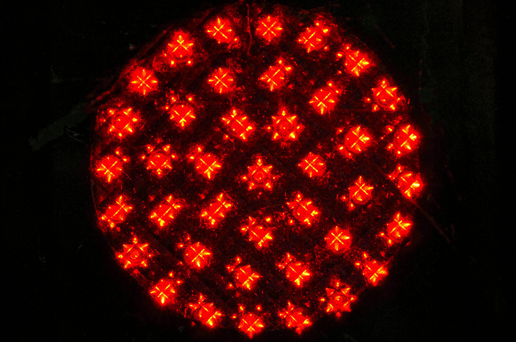 Glühende Geometrie: Das Herz der roten Ampel | Dieses Foto fängt die technologische Ästhetik einer roten Ampel aus nächster Nähe ein. Anstelle einer homogenen Leuchtfläche offenbart der Fokus eine präzise Anordnung einzelner LED-Cluster. Die runden Lichtpunkte sind in einem rautenförmigen Gitter angeordnet und strahlen ein intensives, sattes Rot aus. Durch die Lichtbrechung entstehen kleine, kreuzförmige Strahlen um die einzelnen Lichtquellen, was der Aufnahme eine beinahe feurige oder sternenähnliche Dynamik verleiht. Der tiefschwarze Hintergrund lässt die Leuchtkraft der Dioden besonders stark hervortreten und betont die kreisförmige Gesamtstruktur des Ampelsignals. - Realisiert mit Pictrs.com