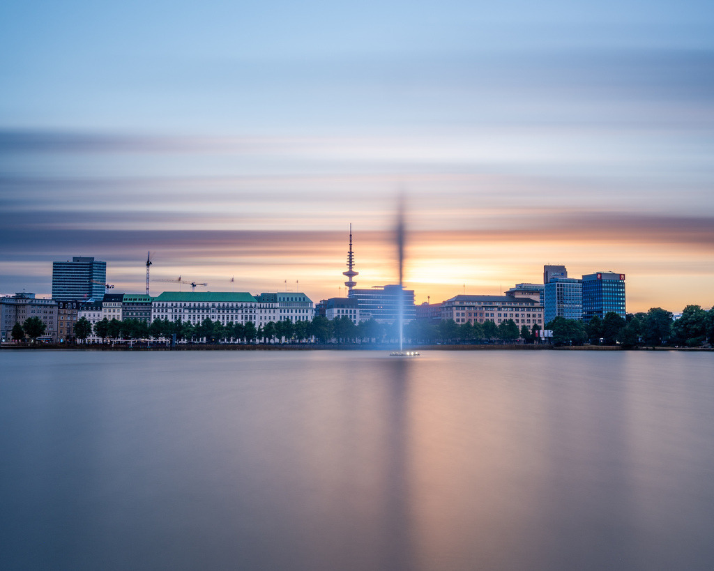 Hamburger Stadtsilhouette im Abendlicht | Eine ruhige Langzeitbelichtung der Hamburger Innenstadt am Wasser. Die sanften Farbtöne des Abendhimmels und die weich gezeichnete Wasseroberfläche verleihen der Stadtsilhouette eine minimalistische und atmosphärische Wirkung. - Realisiert mit Pictrs.com