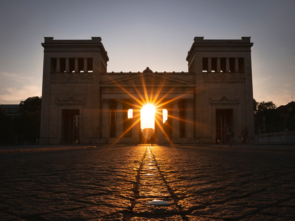 Lichtblick am Königsplatz | Die untergehende Sonne steht perfekt zwischen den Säulen der Propyläen am Münchner Königsplatz und strahlt wie ein leuchtender Stern in den Abend hinein. Die Szenerie wirkt fast symbolisch – als würde das Licht einen Weg durch die Geschichte bahnen. Die tiefstehende Perspektive auf das historische Bauwerk unterstreicht die Strahlkraft dieses Moments und fängt die besondere Atmosphäre dieses Sommerabends ein. - Realisiert mit Pictrs.com
