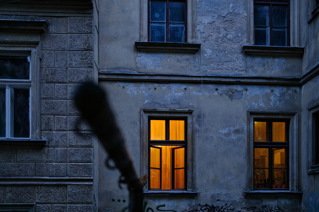 Wohnhaus mit beleuchtetem Fenster am Abend | Ljubljana, Slowenien - June 22, 2025: Wohnhaus mit beleuchtetem Fenster am Abend, im Vordergrund eine Teppichstange. - Realisiert mit Pictrs.com
