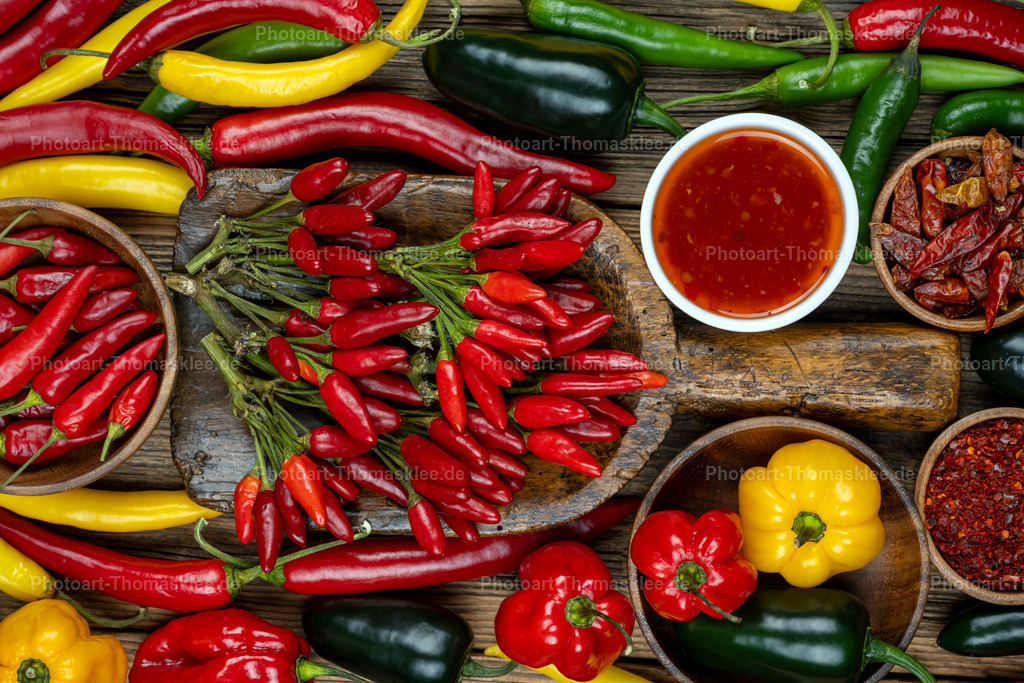 Bunte Chili- und Paprika-Vielfalt auf rustikalem Holz | Zahlreiche Chili- und Paprikasorten bilden ein farbenfrohes Mosaik auf verwittertem Holzuntergrund. Leuchtende rote, gelbe und grüne Schoten ergänzen runde Paprika in verschiedenen Größen. Ein traditionelles Holzbrett trägt einen dekorativen Strauß kleiner roter Chilischoten mit grünen Stielen. Holzschalen präsentieren weitere Chilisorten, während eine Porzellanschale rote Sauce bereithält. Diese kunstvolle Food-Fotografie kombiniert natürliche Holztexturen mit der lebendigen Farbpalette frischer Gewürze und inspiriert zu würzigen Kochabenteuern. - Realisiert mit Pictrs.com