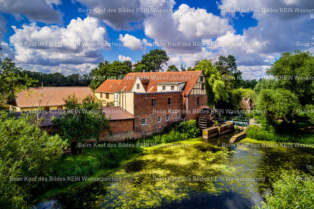 Wassermühle Elbeu bei Wolmirstedt-0005 | Das Luftbild zeigt die historische Mühle in Elbeu aus einer Drohnenperspektive an einem sonnigen Sommertag. Im Vordergrund liegt eine ruhige Wasserfläche, während über der Mühle markante Quellwolken den Himmel prägen und der Aufnahme eine eindrucksvolle Tiefe verleihen. Die farbenfrohe Landschaft bildet einen harmonischen Rahmen für das technische Denkmal.Die Mühle Elbeu steht beispielhaft für die Entwicklung der Getreideverarbeitung vom handwerklichen Mühlenbetrieb zur industriellen Produktion. Im Laufe ihrer Geschichte wurde die Antriebstechnik mehrfach modernisiert – von der ursprünglichen Wind- beziehungsweise Wasserkraft bis hin zum elektrischen Antrieb. Walzenstühle, Elevatoren und Förderanlagen ersetzten nach und nach die traditionellen Mahlgänge und steigerten die Leistungsfähigkeit der Mühle erheblich. Solche Mühlen konnten täglich mehrere Tonnen Getreide zu Mehl und Futtermitteln verarbeiten und waren über Jahrzehnte ein wichtiger Bestandteil der regionalen Landwirtschaft.Die Drohnenaufnahme verbindet historische Mühlentechnik mit der sommerlichen Landschaft der Elbaue. Die markanten Quellwolken, das Wasser im Vordergrund und die eindrucksvolle Architektur der Mühle machen das Bild zu einer gelungenen Verbindung aus Technik, Landschaft und Industriegeschichte. - Realisiert mit Pictrs.com