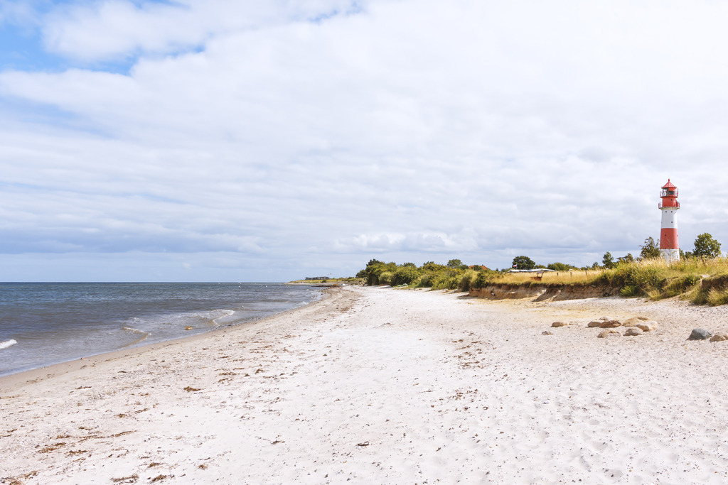 Wandbild: Strandidylle Falshöft - Meeresweite und Leuchtturmblick | Dieses helle, weitläufige Wandbild holt das Gefühl unbeschwerter Küstenstunden direkt in Ihre Räume. Der weiße Sandstrand führt den Blick sanft entlang des ruhigen Meeres bis zum rot-weißen Leuchtturm Falshöft, der prägnant am Horizont emporragt. Unter einem weichen Wolkenhimmel entfaltet das Motiv eine befreiende Klarheit und zeitlose Eleganz – eine edle Bereicherung für moderne, stilvolle Wohn- und Arbeitswelten mit Gespür für maritime Ästhetik.