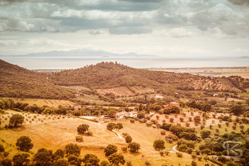 Panoramablick von Scarlino auf die Maremma und das Meer | Ein weitläufiger Panoramablick von der mittelalterlichen Stadt Scarlino in der Toskana auf die umliegende Maremma-Landschaft und das Tyrrhenische Meer. Das Bild zeigt die charakteristischen Hügel mit Olivenhainen, goldenen Feldern und die Küste in der Ferne, möglicherweise mit Blick auf die Insel Elba. Es fängt die warme und historische Atmosphäre der Region ein. - Realisiert mit Pictrs.com